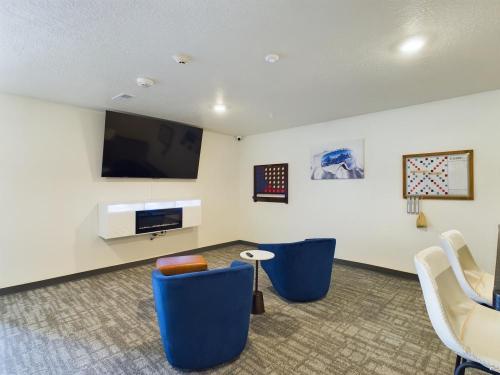 Apartments in Clearfield UT Room with a wall-mounted TV, two blue chairs, a small table, wall games, and a shelf with games. Neutral carpet and walls with minimal decoration.