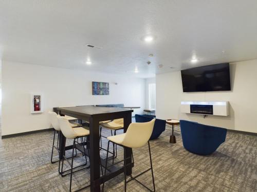 Apartments in Clearfield UT Modern lounge area with a high table, chairs, blue armchairs, a wall-mounted TV, and a decorative fireplace beneath it. Carpeted floor and white walls with a colorful wall art piece.