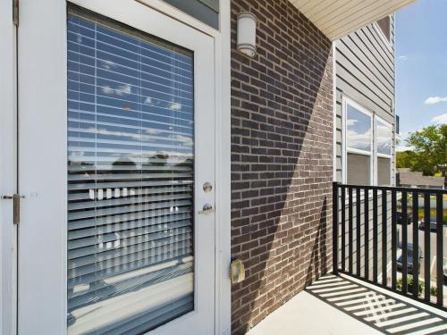 Apartments in Clearfield UT Small balcony with a white door and blinds, brick wall, and black railing. Sunlight casts shadows on the floor. Clear sky and trees visible in the distance.