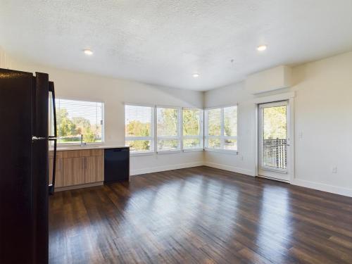 Apartments in Clearfield UT A vacant room with wooden flooring, a row of large windows, and a glass door leading to a small balcony. There's a black refrigerator and dishwasher next to a wooden countertop.