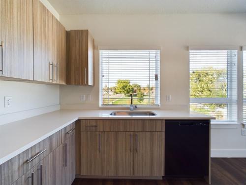 Apartments in Clearfield UT Modern kitchen with wood cabinets, white countertops, a black dishwasher, and a stainless steel sink under two windows with blinds, overlooking a green outdoor area.