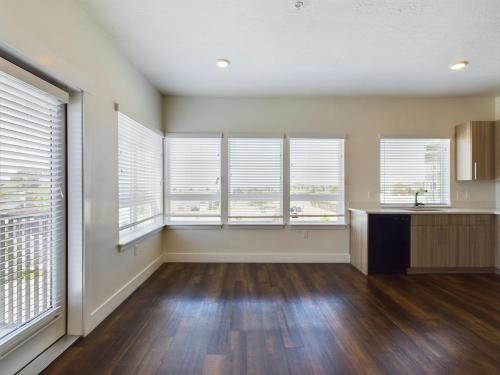 Apartments in Clearfield UT Empty room with large windows, wood flooring, and a small kitchen area. White blinds cover the windows.