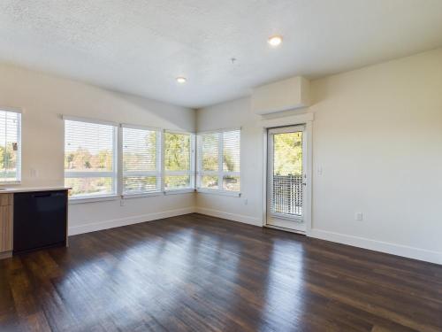 Apartments in Clearfield UT Empty room with light wood flooring, large windows with blinds, and a door leading to a balcony. Walls are painted white and ceiling lights are on.